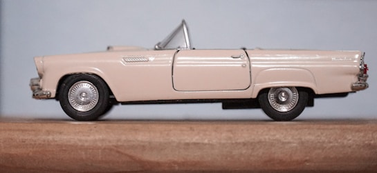 A model of a vintage convertible car with a beige exterior is positioned on a wooden surface. The car features detailed silver wheel caps, a prominent grille, and a sleek, classic design.