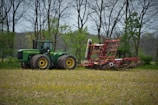 A large green tractor is parked in a field, attached to a red agricultural implement, presumably a plow or harrow. The field appears recently plowed or prepared for planting, and it's surrounded by tall, bare trees with some green foliage indicating spring. The sky is overcast, providing diffused lighting across the scene.
