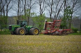 A large green tractor is parked in a field, attached to a red agricultural implement, presumably a plow or harrow. The field appears recently plowed or prepared for planting, and it's surrounded by tall, bare trees with some green foliage indicating spring. The sky is overcast, providing diffused lighting across the scene.
