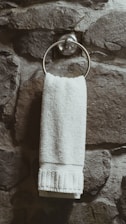 A white towel hangs neatly from a metallic ring attached to a textured stone wall. The wall has a rugged and natural appearance with varying shades of gray and brown.