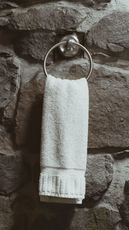 A white towel hangs neatly from a metallic ring attached to a textured stone wall. The wall has a rugged and natural appearance with varying shades of gray and brown.