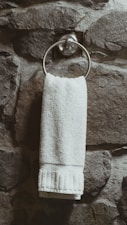 A white towel hangs neatly from a metallic ring attached to a textured stone wall. The wall has a rugged and natural appearance with varying shades of gray and brown.