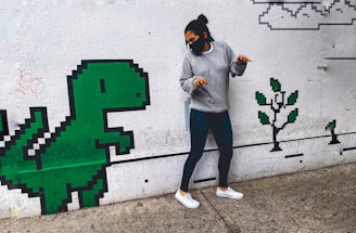 A person wearing a grey sweater, jeans, and a black face mask is playfully posing next to a wall mural featuring a pixelated green dinosaur and a pixel tree. The mural has a retro, 8-bit video game style, with simple shapes and blocks.
