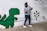 A person wearing a grey sweater, jeans, and a black face mask is playfully posing next to a wall mural featuring a pixelated green dinosaur and a pixel tree. The mural has a retro, 8-bit video game style, with simple shapes and blocks.