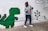 A person wearing a grey sweater, jeans, and a black face mask is playfully posing next to a wall mural featuring a pixelated green dinosaur and a pixel tree. The mural has a retro, 8-bit video game style, with simple shapes and blocks.