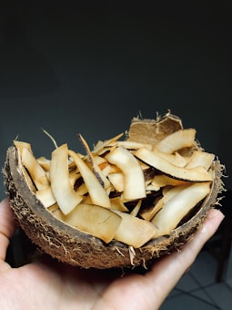 A hand holding a halved coconut shell filled with thinly sliced, toasted coconut chips. The shell is rough and textured, with fibers visible on the outside. The background is dark, enhancing the focus on the coconut.