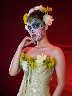 A person with elaborate sugar skull makeup, typical of Dia de los Muertos celebrations, featuring ornate designs around the eyes and lips. The individual is wearing a strapless, patterned dress adorned with large, colorful flowers. Their hair is styled with a crown of yellow and white flowers, and they pose thoughtfully against a deep red background.