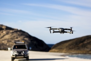A compact drone hovering over a sandy beach under clear blue skies.