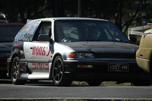 A modified car with a prominent 'Canon' decal on the side panel is parked on the road. The car appears to be an older model hatchback with custom rims and a unique paint job. The surroundings include other vehicles in close proximity, partially visible, highlighting an automotive setting, possibly at an event or meetup.