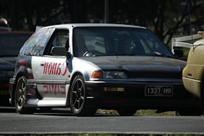 A modified car with a prominent 'Canon' decal on the side panel is parked on the road. The car appears to be an older model hatchback with custom rims and a unique paint job. The surroundings include other vehicles in close proximity, partially visible, highlighting an automotive setting, possibly at an event or meetup.
