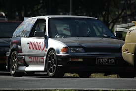A modified car with a prominent 'Canon' decal on the side panel is parked on the road. The car appears to be an older model hatchback with custom rims and a unique paint job. The surroundings include other vehicles in close proximity, partially visible, highlighting an automotive setting, possibly at an event or meetup.