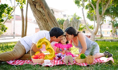 A family picnic scene with a young child between two adults who are kissing the child on the cheeks. They are sitting on a red and white checkered blanket on the grass, surrounded by a variety of fruits including a pineapple, watermelon, and limes. A golden balloon in the shape of the number 3 is beside them, suggesting a celebration. The setting is outdoors in a park with trees around, creating a bright and cheerful atmosphere.