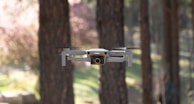 A small white drone hovers in mid-air in a wooded area, with blurred trees and greenery in the background. The drone's design is sleek and compact, equipped with a camera at the front.