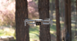 A small white drone hovers in mid-air in a wooded area, with blurred trees and greenery in the background. The drone's design is sleek and compact, equipped with a camera at the front.