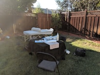 A backyard setup features a massage table with a rolled-up towel and a covered table with various items including a bottle of lotion. Two chairs are placed around the table, and the setting is enclosed by a wooden fence with a tree providing some shade.