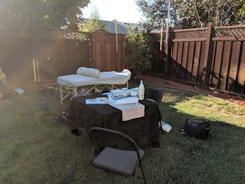 A backyard setup features a massage table with a rolled-up towel and a covered table with various items including a bottle of lotion. Two chairs are placed around the table, and the setting is enclosed by a wooden fence with a tree providing some shade.