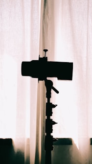 A silhouette of a camera on a tripod is positioned in front of a curtain, with soft natural light filtering through, creating a minimalistic and artistic effect.