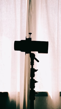 A silhouette of a camera on a tripod is positioned in front of a curtain, with soft natural light filtering through, creating a minimalistic and artistic effect.