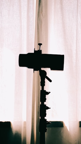 A silhouette of a camera on a tripod is positioned in front of a curtain, with soft natural light filtering through, creating a minimalistic and artistic effect.