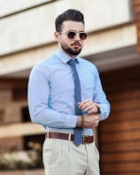 A man standing confidently outdoors, wearing a light blue dress shirt with thin vertical stripes, a blue polka dot tie, and beige trousers. He has a well-groomed beard, slicked-back hair, and is wearing sunglasses. A brown leather belt and wristwatch complete his attire. The background is slightly blurred, hinting at an urban environment.