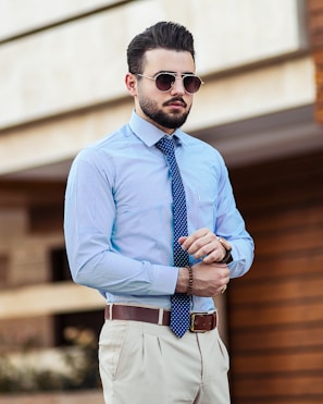 A man standing confidently outdoors, wearing a light blue dress shirt with thin vertical stripes, a blue polka dot tie, and beige trousers. He has a well-groomed beard, slicked-back hair, and is wearing sunglasses. A brown leather belt and wristwatch complete his attire. The background is slightly blurred, hinting at an urban environment.