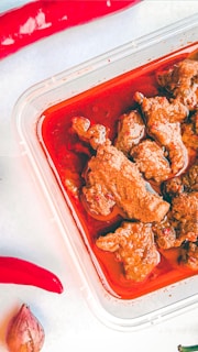 A plastic container holds pieces of meat in a rich, red sauce. Surrounding the container are two red chili peppers and a small shallot on a light surface.