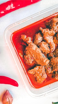 A plastic container holds pieces of meat in a rich, red sauce. Surrounding the container are two red chili peppers and a small shallot on a light surface.