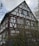 A traditional half-timbered house with white walls and dark brown wooden beams. The structure features multiple windows with green shutters. Surrounding the house is dense greenery and shrubbery, while the sky above is partly cloudy.