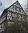 A traditional half-timbered house with white walls and dark brown wooden beams. The structure features multiple windows with green shutters. Surrounding the house is dense greenery and shrubbery, while the sky above is partly cloudy.