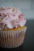 A cupcake with light pink frosting adorned with decorative sprinkles, including small white stars and pink and white spherical beads. The cupcake has a textured paper wrapper and is placed on a dark surface, creating a contrast that highlights the delicate details of the frosting.