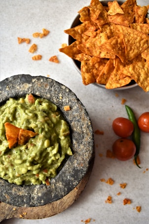 A bowl of guacamole is placed on a rustic stone surface, garnished with a tortilla chip. Beside it, a bowl is filled with crispy, golden tortilla chips. Scattered around are pieces of broken chips, along with fresh tomatoes and a green chili pepper on a light gray background.