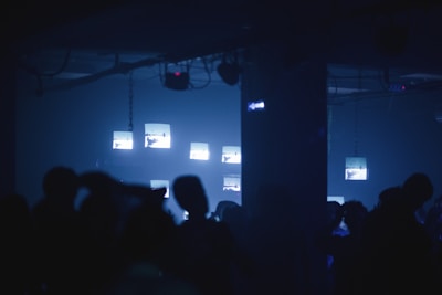 A dimly lit room with several illuminated screens suspended from the ceiling. Silhouettes of people are visible in the foreground, suggesting a club or concert setting. The atmosphere is dark and shadowy with a modern, tech-inspired vibe.