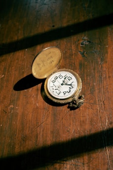 An antique brass pocket compass lies open on a wooden surface, with sunlight casting shadows. The compass lid is engraved and the dial shows direction markings.