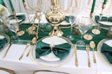 A luxurious table setting featuring a white tablecloth with gold-rimmed plates and green satin napkins. Golden cutlery and tall, elegant glassware are arranged symmetrically. Green candles in gold holders and decorative gold objects enhance the opulent appearance.