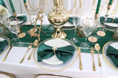 A luxurious table setting featuring a white tablecloth with gold-rimmed plates and green satin napkins. Golden cutlery and tall, elegant glassware are arranged symmetrically. Green candles in gold holders and decorative gold objects enhance the opulent appearance.