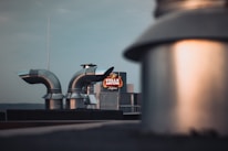 Industrial rooftop with metal ventilation ducts and a blurred foreground. A brightly illuminated Stella Artois sign is visible in the background against a clear sky.