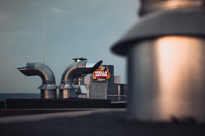 Industrial rooftop with metal ventilation ducts and a blurred foreground. A brightly illuminated Stella Artois sign is visible in the background against a clear sky.