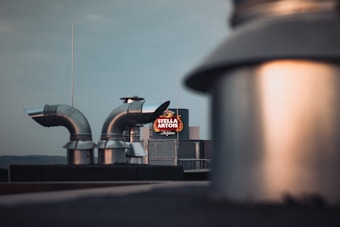 Industrial rooftop with metal ventilation ducts and a blurred foreground. A brightly illuminated Stella Artois sign is visible in the background against a clear sky.