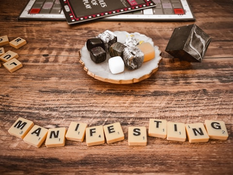 A wooden table displays Scrabble tiles spelling the word 'MANIFESTING.' Nearby, a decorative tray holds a variety of stones, including obsidian, white howlite, and tumbled stones. A black geometric object and part of a Monopoly board game are visible in the background, creating a juxtaposition of game elements and spiritual decor.