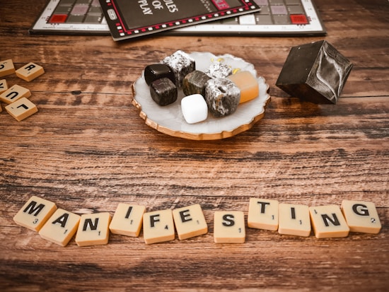 A wooden table displays Scrabble tiles spelling the word 'MANIFESTING.' Nearby, a decorative tray holds a variety of stones, including obsidian, white howlite, and tumbled stones. A black geometric object and part of a Monopoly board game are visible in the background, creating a juxtaposition of game elements and spiritual decor.