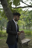A man in a suit stands outdoors leaning against a tree. He appears to be adjusting his wristwatch while looking attentively to his right. The setting is a lush, green park with a wooden bench and trees in the background, creating a calm and natural atmosphere.