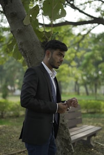 A man in a suit stands outdoors leaning against a tree. He appears to be adjusting his wristwatch while looking attentively to his right. The setting is a lush, green park with a wooden bench and trees in the background, creating a calm and natural atmosphere.