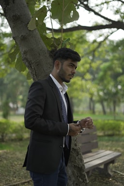 A man in a suit stands outdoors leaning against a tree. He appears to be adjusting his wristwatch while looking attentively to his right. The setting is a lush, green park with a wooden bench and trees in the background, creating a calm and natural atmosphere.