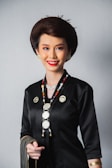 A person dressed in a traditional outfit with intricate jewelry, including a long beaded necklace and gold earrings. The attire is predominantly black with gold accents, and the individual has styled hair and a bright smile.
