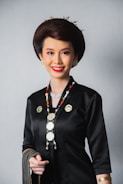A person dressed in a traditional outfit with intricate jewelry, including a long beaded necklace and gold earrings. The attire is predominantly black with gold accents, and the individual has styled hair and a bright smile.