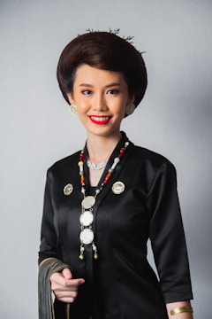 A person dressed in a traditional outfit with intricate jewelry, including a long beaded necklace and gold earrings. The attire is predominantly black with gold accents, and the individual has styled hair and a bright smile.