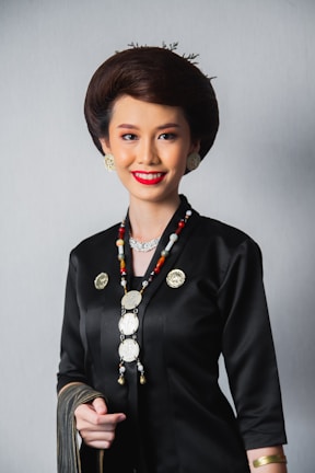 A person dressed in a traditional outfit with intricate jewelry, including a long beaded necklace and gold earrings. The attire is predominantly black with gold accents, and the individual has styled hair and a bright smile.