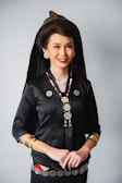 A woman is dressed in traditional attire, featuring a black top adorned with metallic coin decorations and necklaces. She wears a distinct headdress paired with large, round earrings and bracelets on her wrists. Her hair is styled elegantly, complementing her bright red lipstick and warm smile.