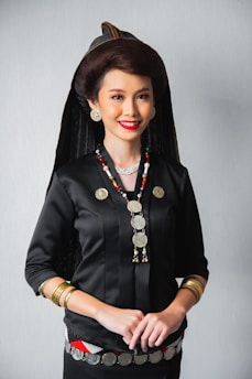 A woman is dressed in traditional attire, featuring a black top adorned with metallic coin decorations and necklaces. She wears a distinct headdress paired with large, round earrings and bracelets on her wrists. Her hair is styled elegantly, complementing her bright red lipstick and warm smile.
