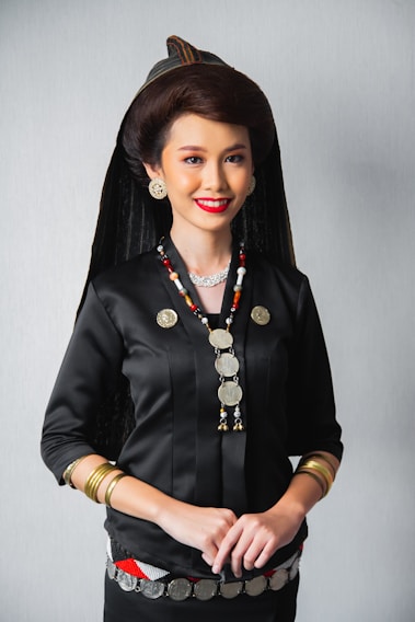 A woman is dressed in traditional attire, featuring a black top adorned with metallic coin decorations and necklaces. She wears a distinct headdress paired with large, round earrings and bracelets on her wrists. Her hair is styled elegantly, complementing her bright red lipstick and warm smile.